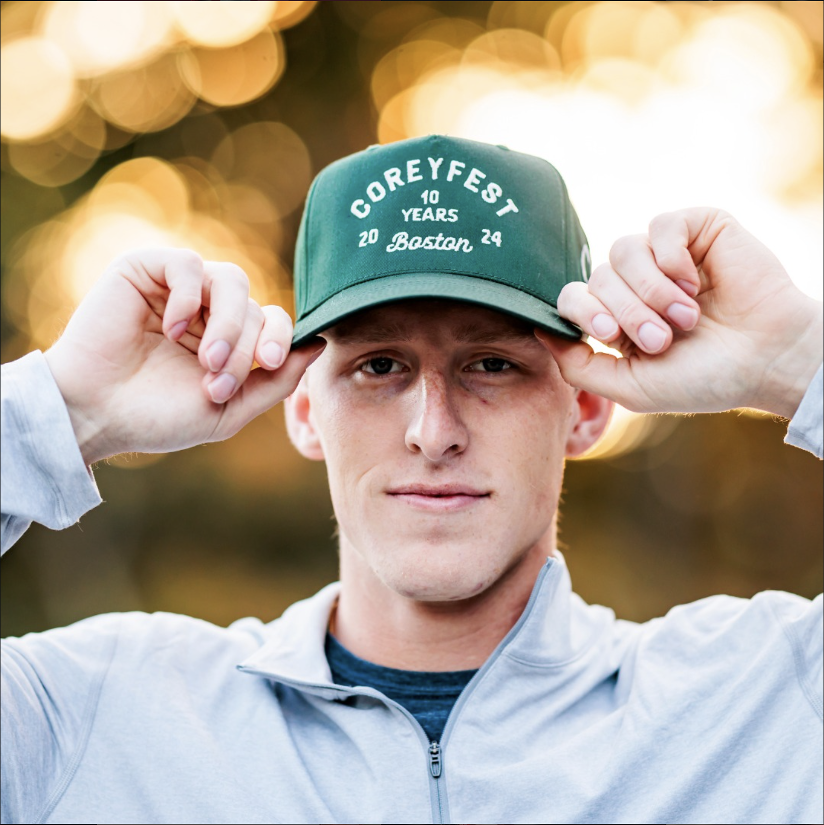 Young man wearing green CoreyFest 10 Years Boston 2024 hat against golden bokeh background