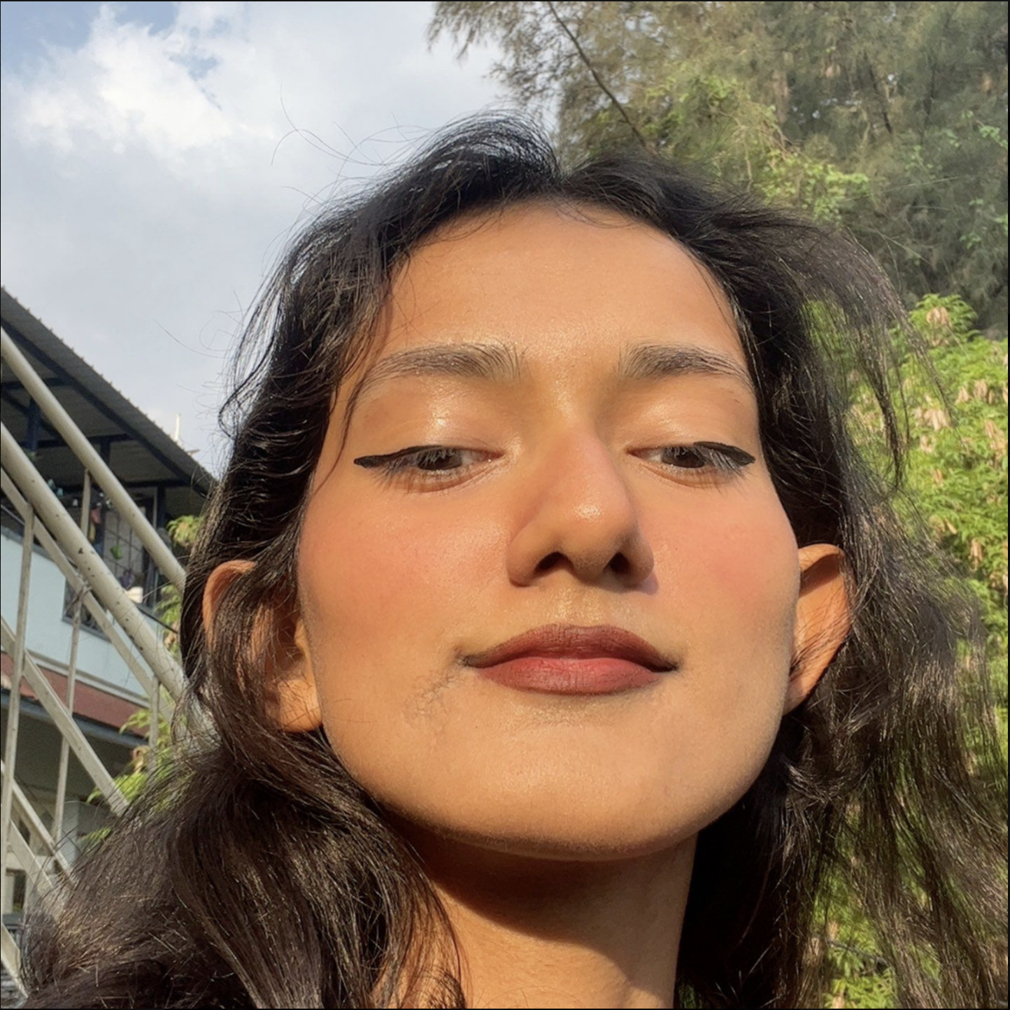 Close-up selfie of Glen, a young woman with dark wavy hair, taken outdoors with trees and a building with stairs in the background