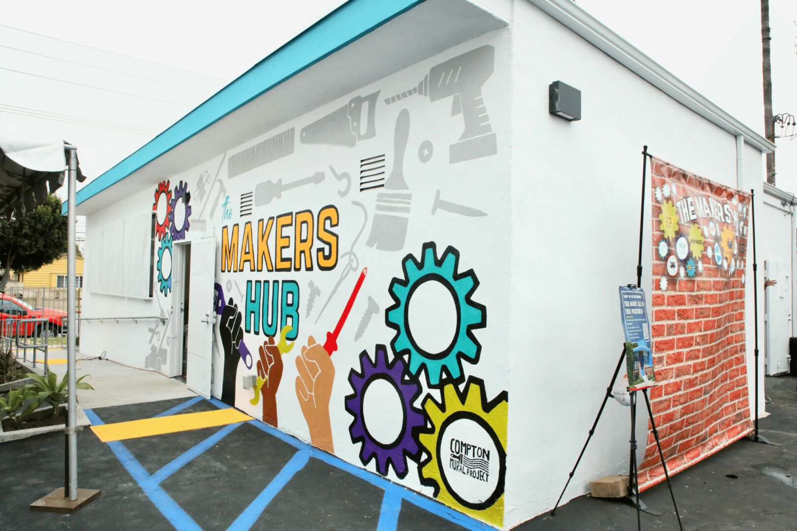The Makers Hub building exterior in Compton featuring a colorful mural with gears, diverse hands holding tools, and the Compton Mural Project logo