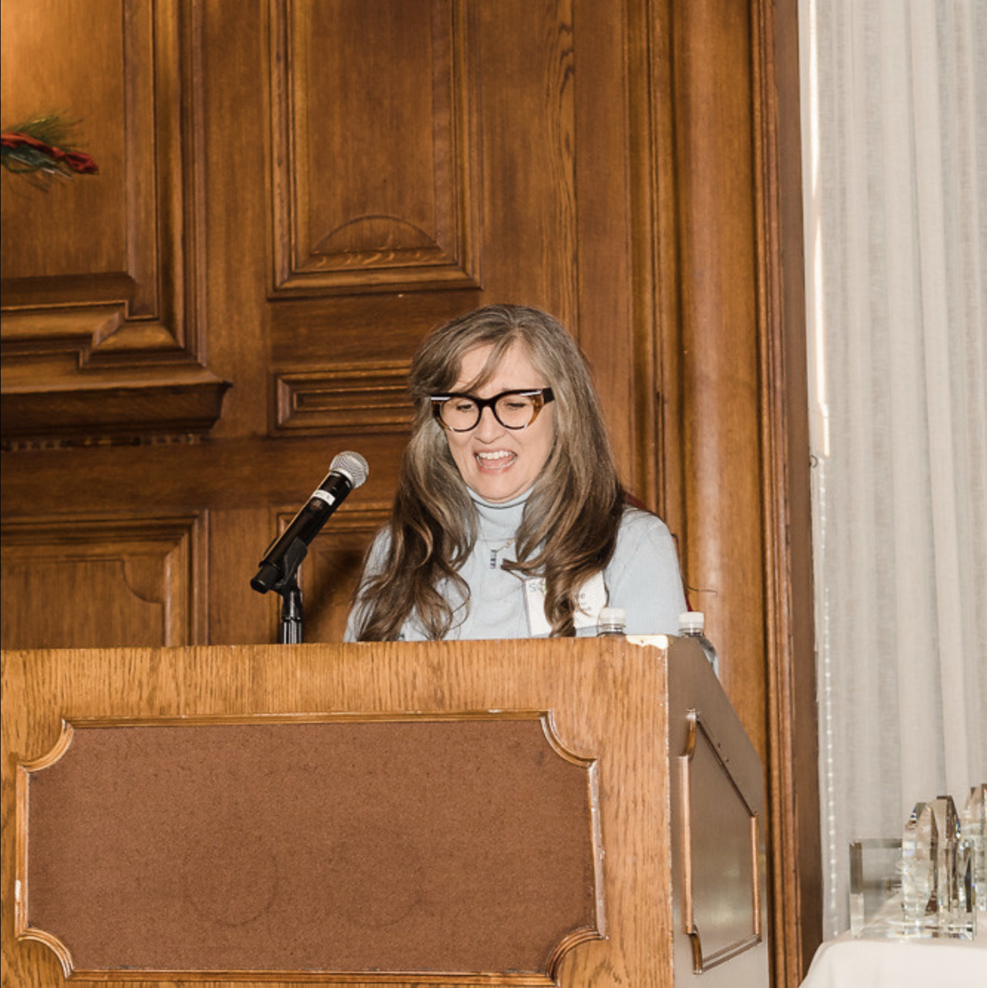 Adrianne Ferree, Executive Director of The Makers Hub, speaking at a podium with microphone in a wood-paneled room
