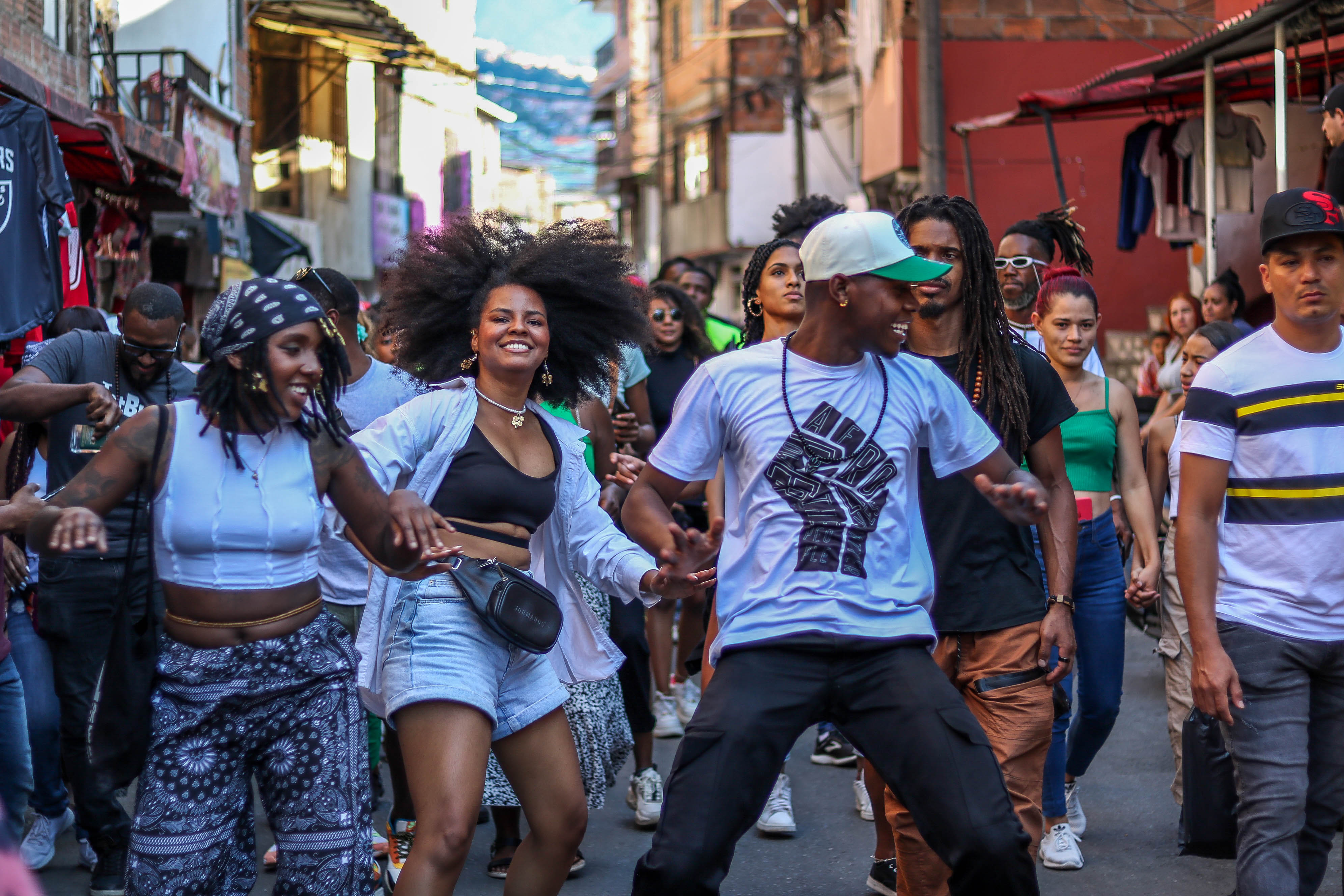 Black Joy Parade street dancing scene in Medellin showing joyful participants dancing together, with a woman with natural afro hair in denim shorts dancing with a man in a white t-shirt and green cap