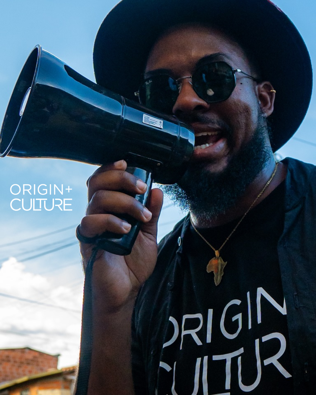 Man with sunglasses and hat speaking into megaphone wearing Origin + Culture t-shirt with Africa pendant necklace